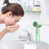 Woman rinsing face next to green ultrasonic face brush in modern bathroom